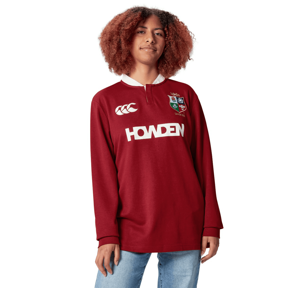 A cheerful person stands with hands by their sides, wearing the red British &amp; Irish Lions 2025 Long Sleeve Classic Jersey by Canterbury, adorned with white logos, smiling against a white background.