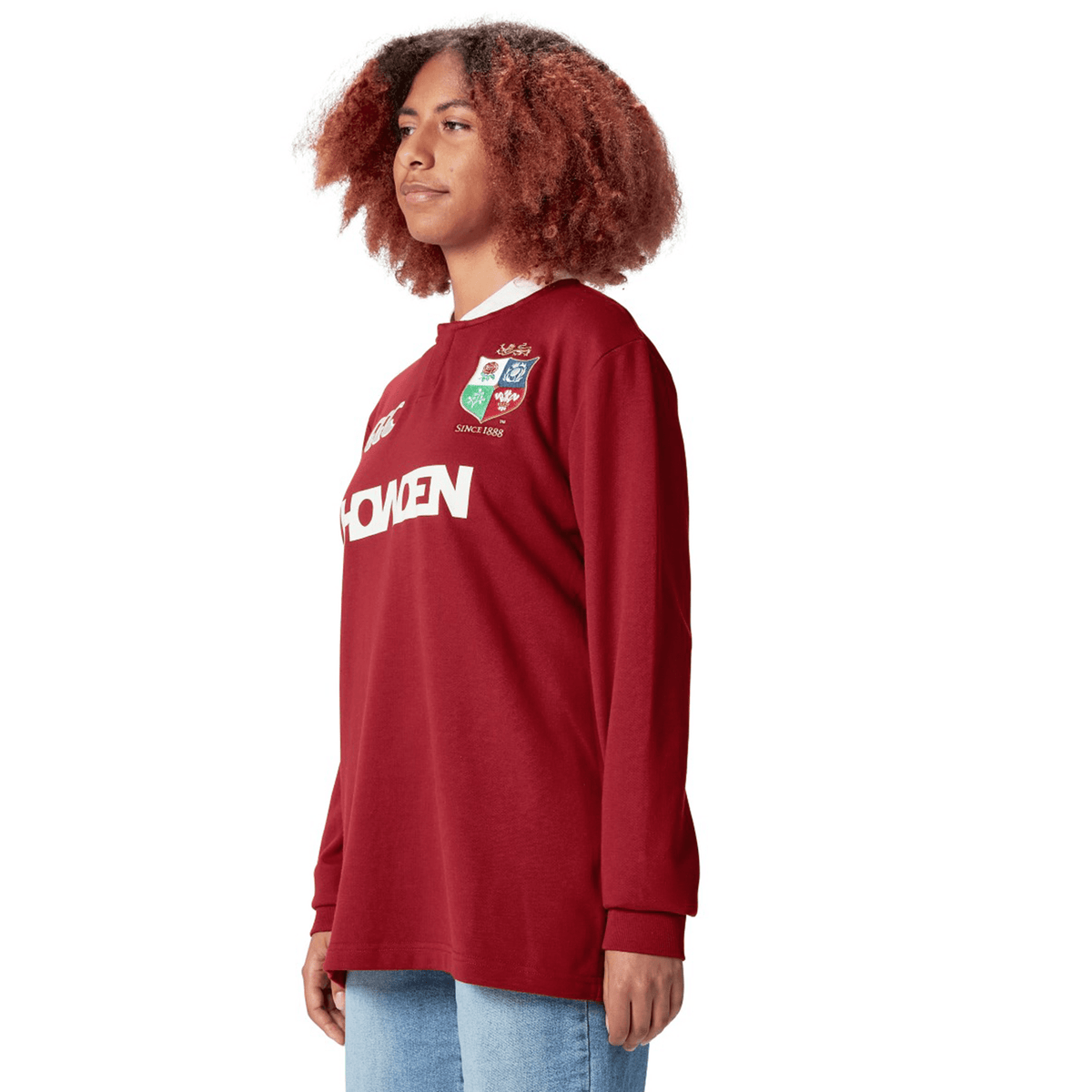 A person stands against a white background, wearing the British &amp; Irish Lions 2025 Long Sleeve Classic Jersey by Canterbury, featuring logos and paired with light blue jeans.