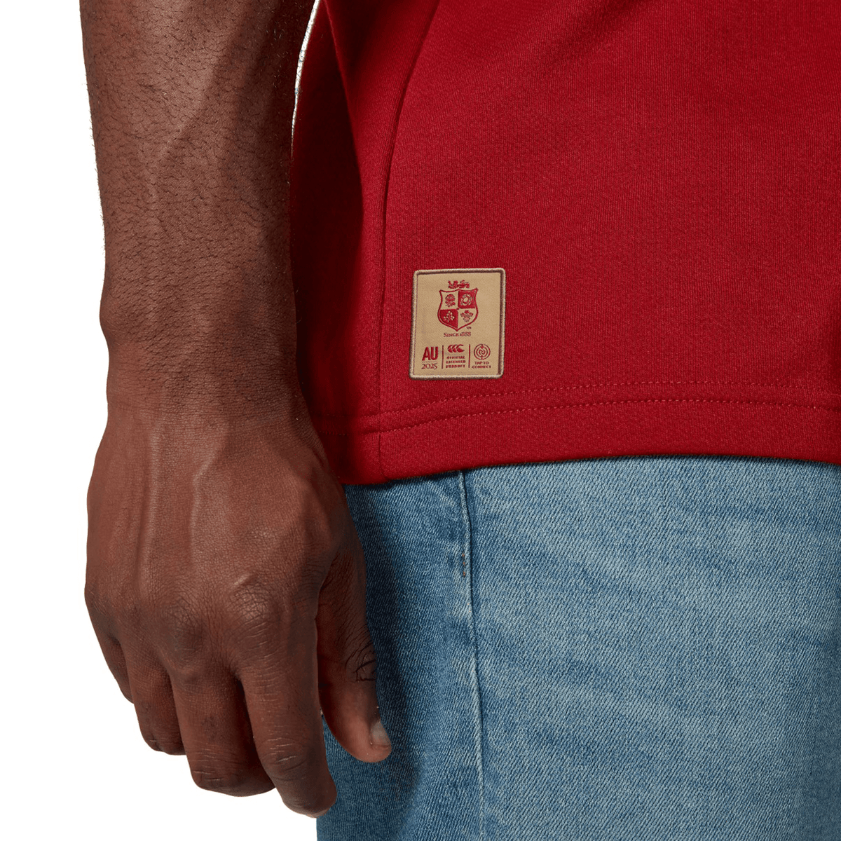 A person in a vibrant British &amp; Irish Lions 2025 Classic Jersey by Canterbury and blue jeans is shown from the waist down, standing with their right arm by their side.