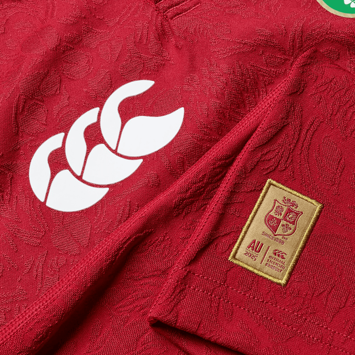 Close-up of a red British &amp; Irish Lions 2025 Youth Players Jersey by Canterbury, crafted from moisture-wicking fabric, showcasing a white logo on the left and a rectangular patch with a shield and text on the right.