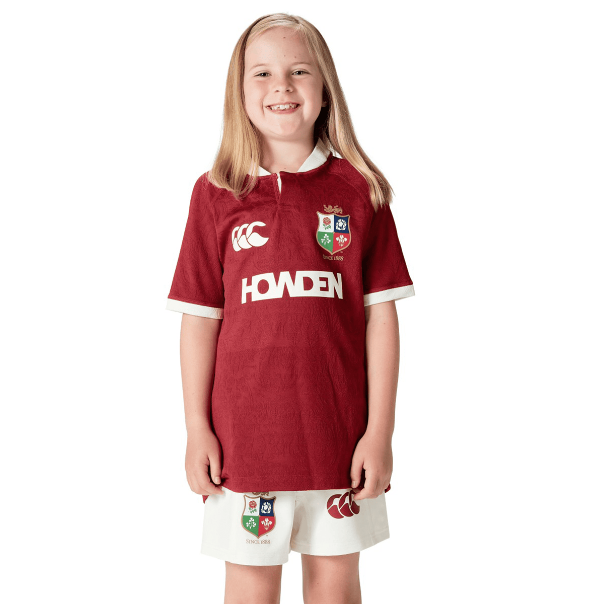 A young girl with long blonde hair smiles, wearing a Youth British &amp; Irish Lions 2025 Players Jersey by Canterbury. The maroon and white rugby jersey and shorts feature logos and moisture-wicking fabric, perfect for active days.