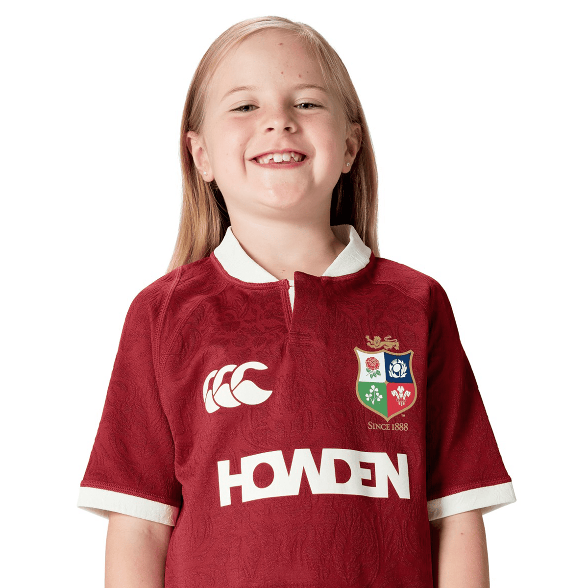 A smiling child proudly wears the maroon Youth British &amp; Irish Lions 2025 Players Jersey by Canterbury, which offers ergonomic paneling for comfort and a moisture-wicking fabric to keep them dry during active play.