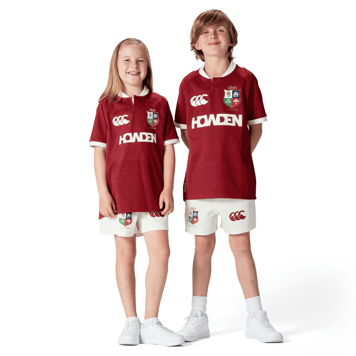 Two smiling children wearing matching red Youth British &amp; Irish Lions 2025 Players Jerseys by Canterbury and white shorts, stand side by side against a plain white background.