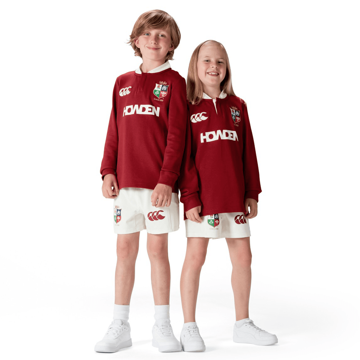 Two children wearing matching Youth British &amp; Irish Lions 2025 Long Sleeve Classic Jerseys by Canterbury in maroon, featuring moisture-wicking fabric and paired with white shorts, stand side by side, smiling.