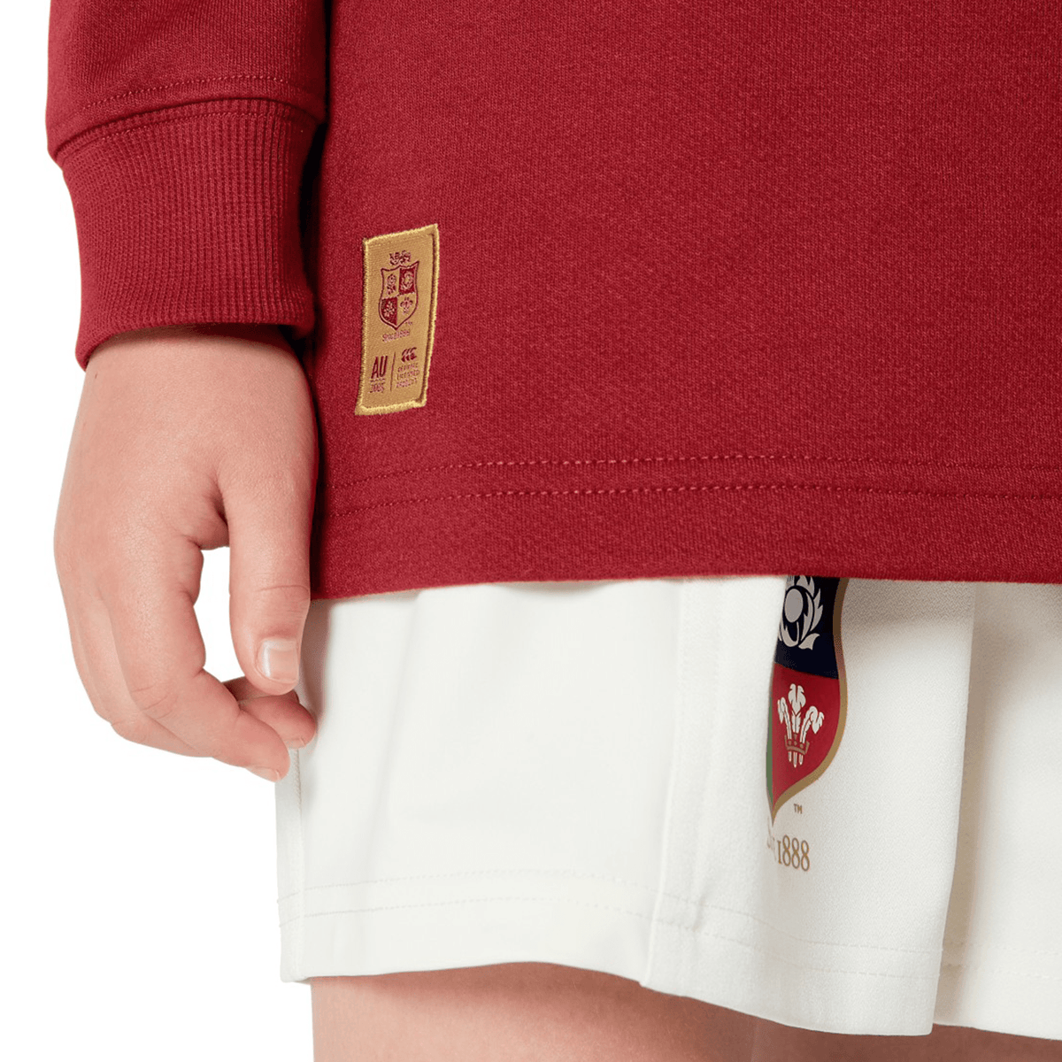 A person models the Youth British &amp; Irish Lions 2025 Long Sleeve Classic Jersey by Canterbury in red with a crest and white shorts, set against a neutral background, highlighting its comfort-driven moisture-wicking fabric.