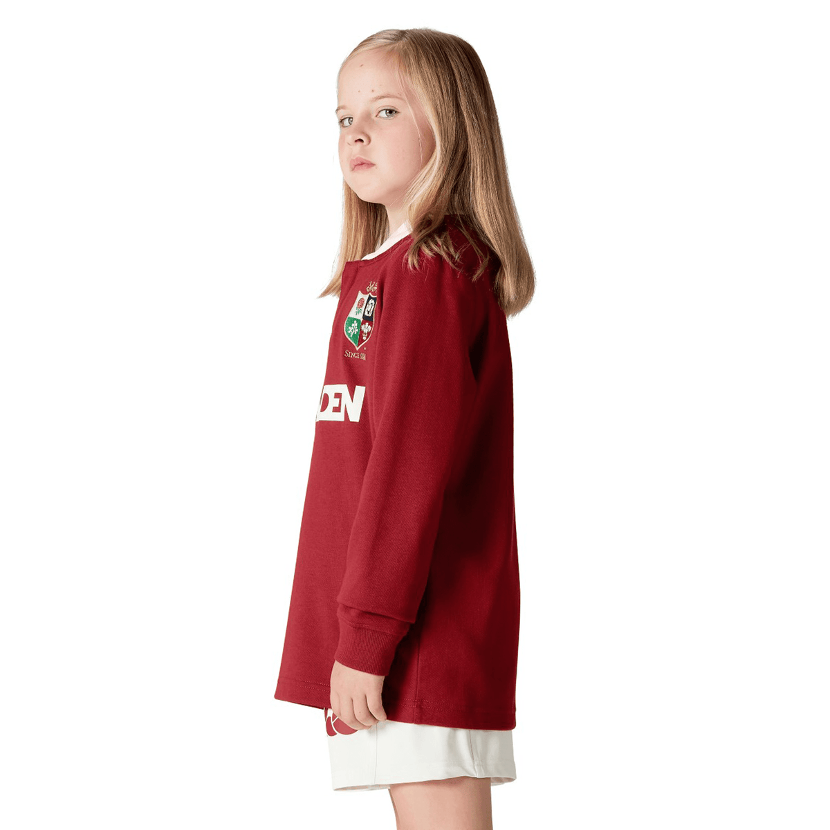 A young girl with long hair stands sideways, wearing the Youth British &amp; Irish Lions 2025 Long Sleeve Classic Jersey by Canterbury and white shorts, looking forward.