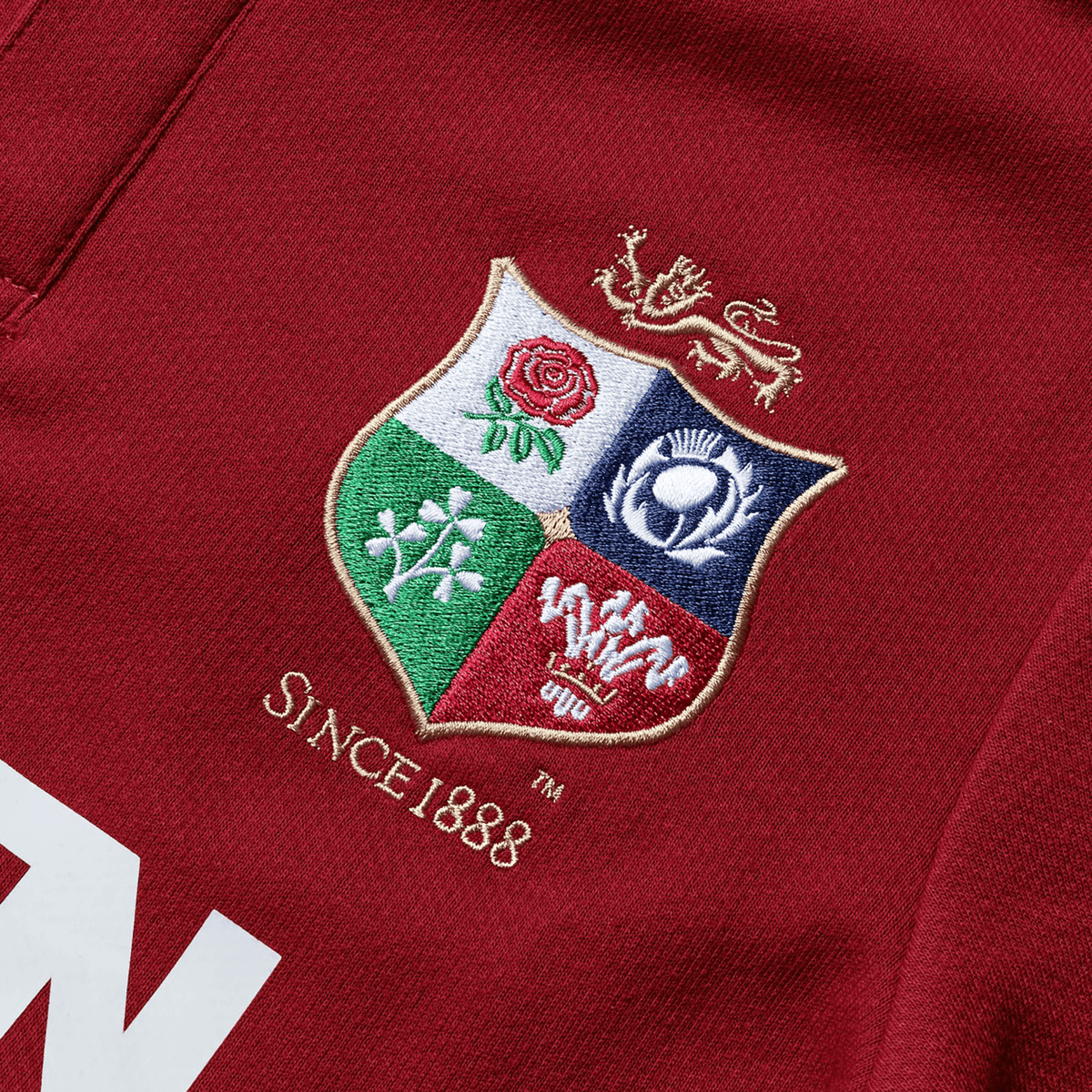 A close-up of the Youth British &amp; Irish Lions 2025 Classic Jersey by Canterbury, featuring a red fabric with an embroidered crest showing a red rose, thistle, shamrock, and dragon. The text "SINCE 1888" honors the legacy of the British &amp; Irish Lions.