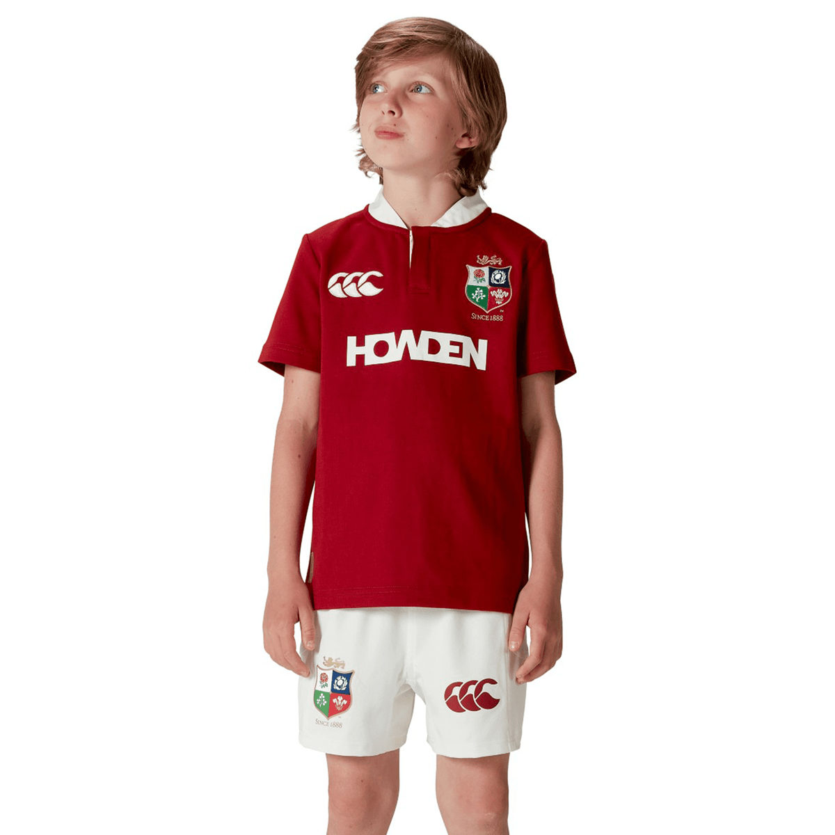 A young boy stands confidently, gazing skyward in a vibrant red Youth British &amp; Irish Lions 2025 Classic Jersey by Canterbury, perfectly paired with logo-adorned white shorts and sleek short sleeves.