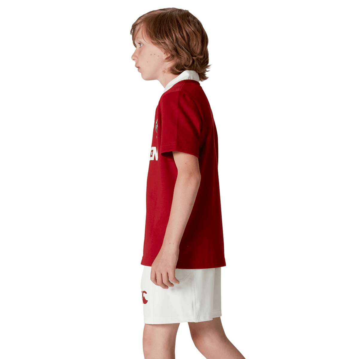 A child with brown hair stands in profile wearing the Youth British &amp; Irish Lions 2025 Classic Jersey by Canterbury in red, paired with white shorts, set against a plain white backdrop.