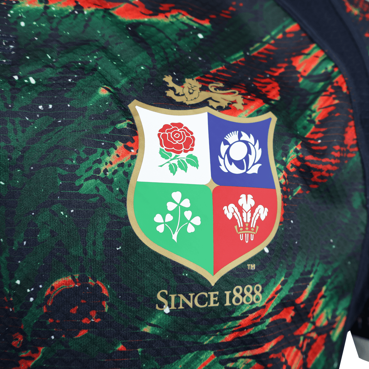 The British &amp; Irish Lions 2025 Replica Warm Up Jersey by Canterbury features an athletic fit with a multicolored crest displaying four symbols and &quot;Since 1888&quot; on a dark, patterned background made from moisture-wicking fabric.
