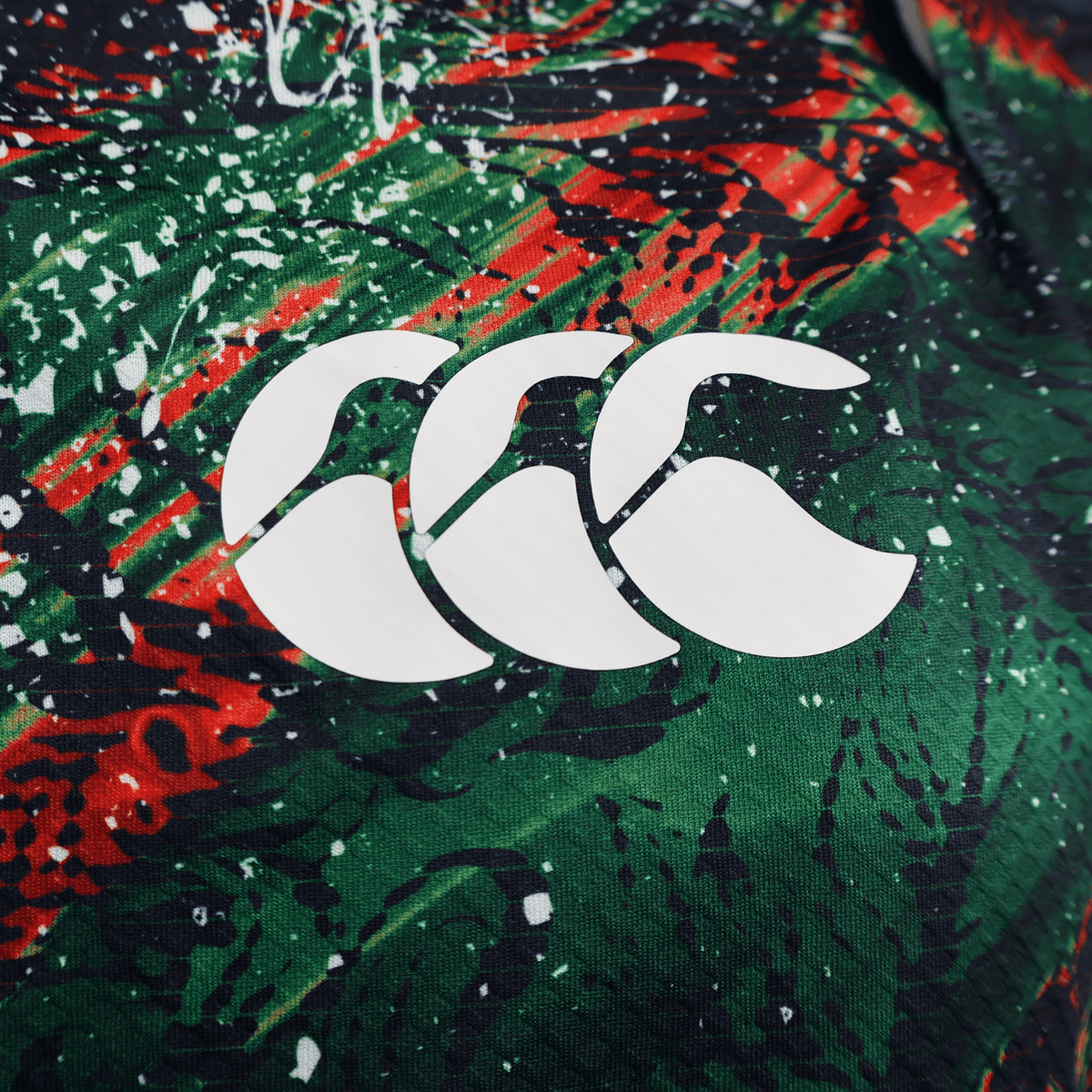 Close-up of the British &amp; Irish Lions 2025 Replica Warm Up Jersey by Canterbury, featuring its red, green, and black pattern. The athletic fit fabric showcases a white logo with three curved shapes, designed for optimal performance with moisture-wicking properties.