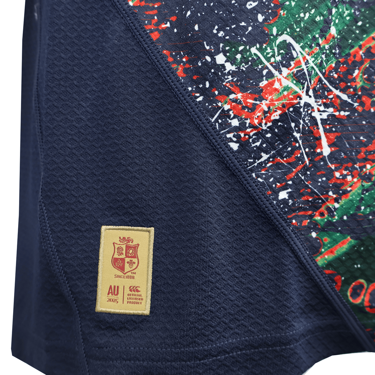 A close-up of the British &amp; Irish Lions 2025 Replica Warm Up Jersey by Canterbury in blue athletic fit, featuring a vibrant splatter design on one side and a stitched patch with crest and text on the other. The moisture-wicking fabric ensures comfort throughout wear.