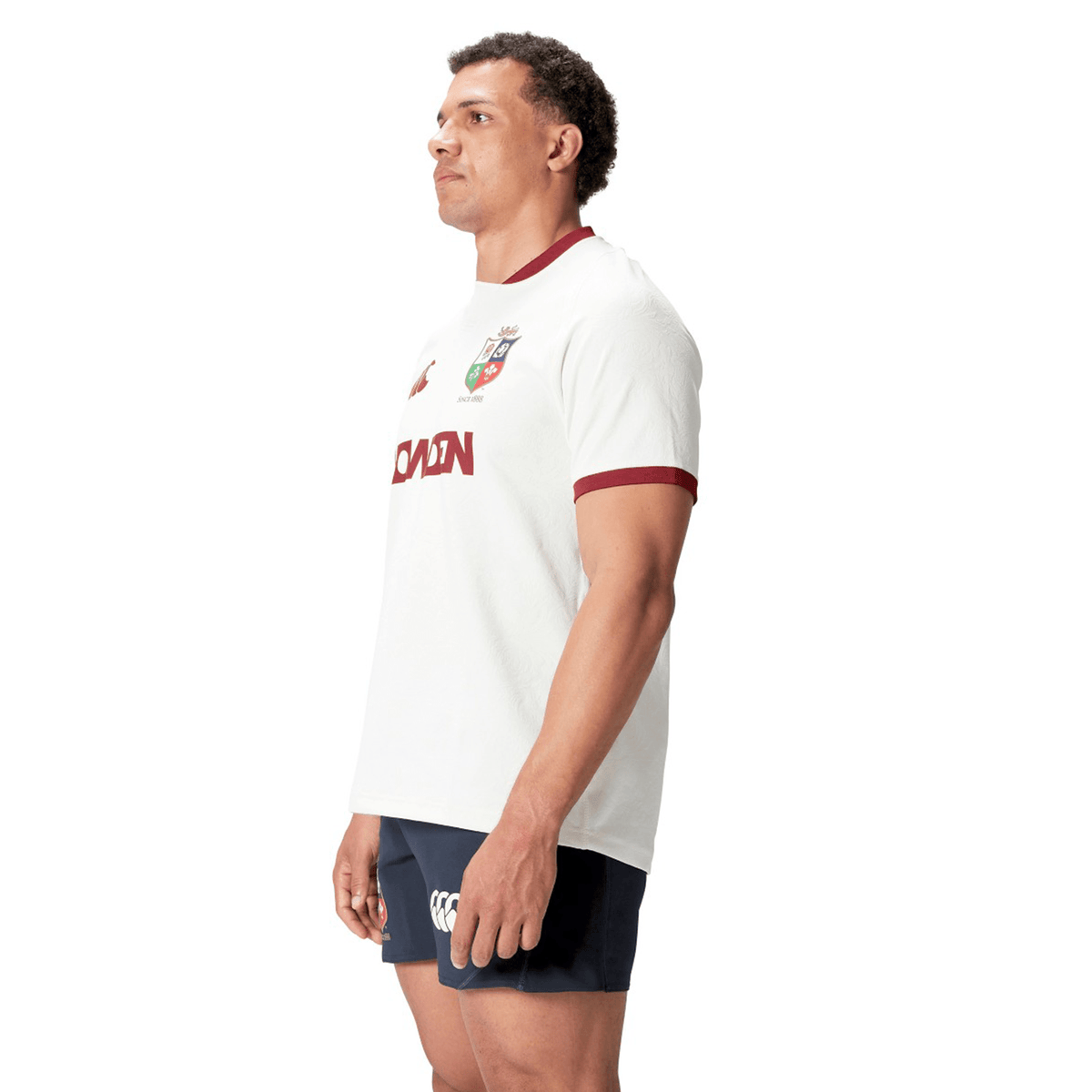 A man wearing the British &amp; Irish Lions 2025 Training Jersey by Canterbury, made from recycled polyester, pairs it with navy shorts as he stands sideways, looking ahead.