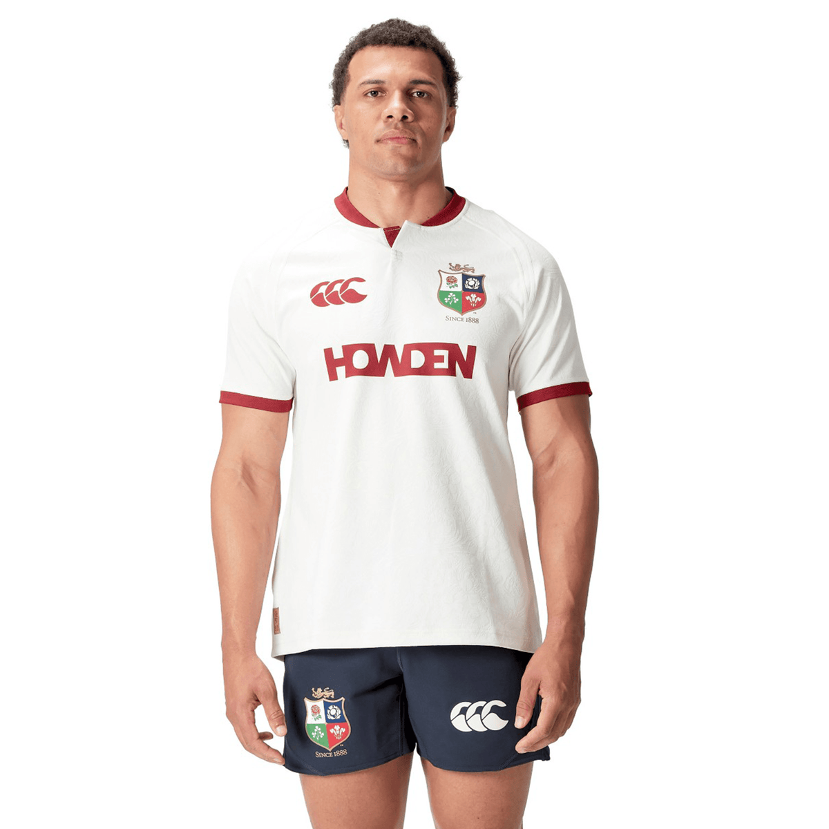 A person stands facing forward wearing a white British &amp; Irish Lions 2025 Training Jersey by Canterbury, made from recycled polyester, paired with navy shorts featuring logos.
