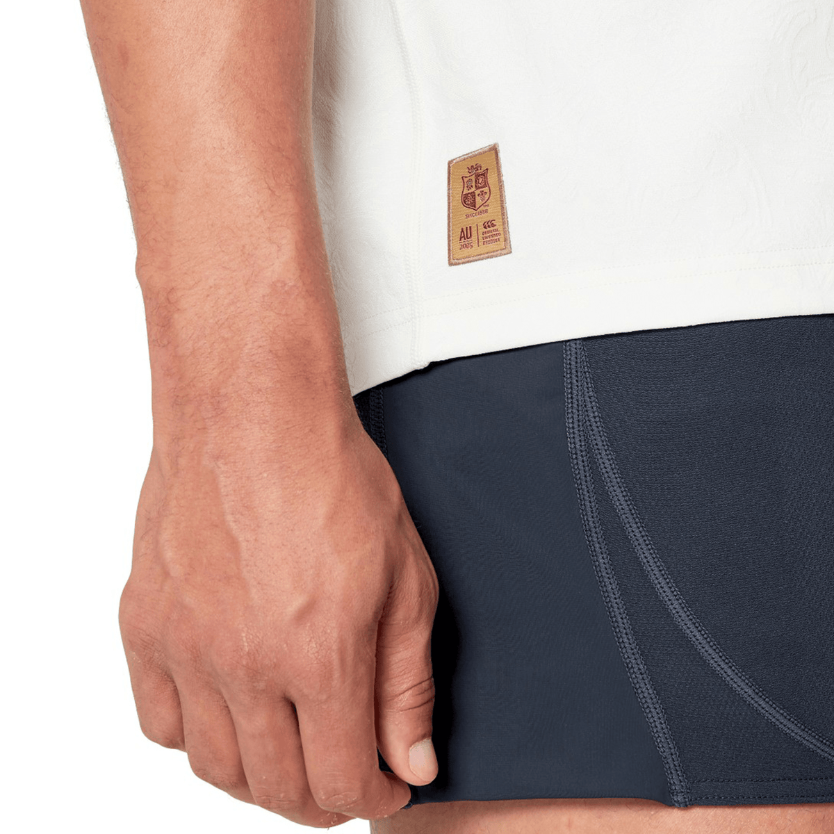 Close-up of a person’s hand resting by their side, wearing a white top with Canterbury branding and dark athletic shorts featuring visible stitching. A small label on the top is reminiscent of the British &amp; Irish Lions 2025 Training Jersey by Canterbury, designed with ergonomically placed panels for comfort.