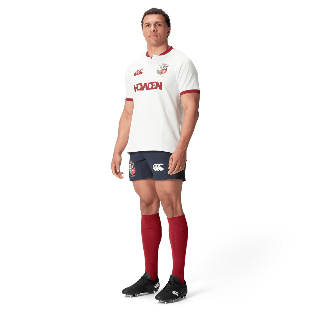 A rugby player wearing the British &amp; Irish Lions 2025 Training Jersey by Canterbury, composed of recycled polyester and featuring a white and red design, stands against a white backdrop, glancing to the side.