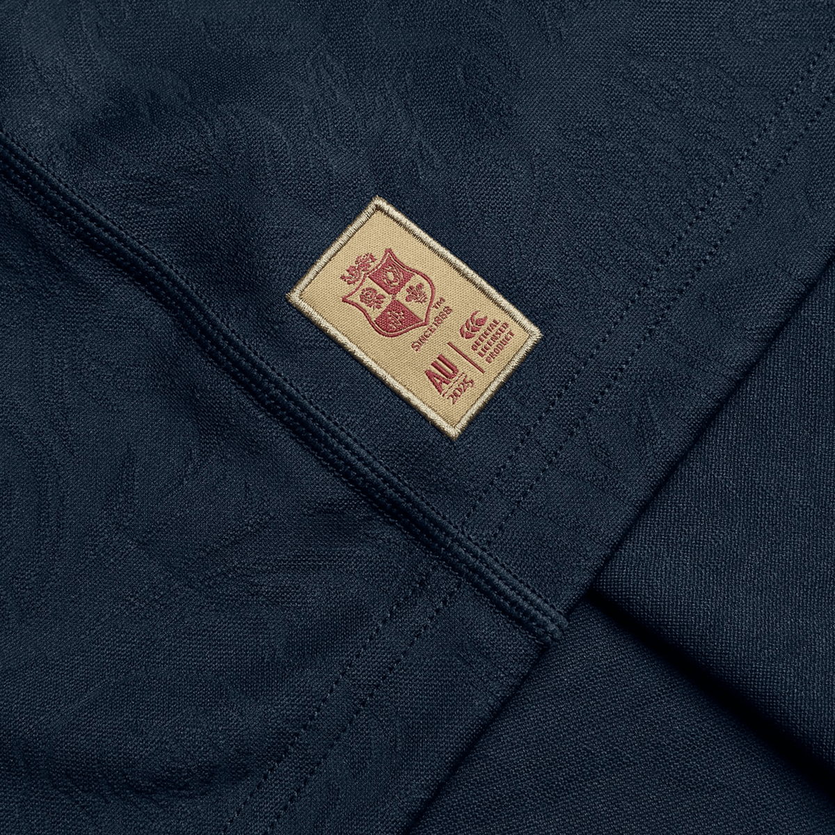 Crafted by Canterbury, the British &amp; Irish Lions 2025 Training Jersey showcases a dark fabric adorned with a beige patch. This patch features a small crest design and text, akin to the iconic replica training jersey. Seamlessly sewn onto the smooth material, it perfectly integrates with strategically placed panels for enhanced comfort.