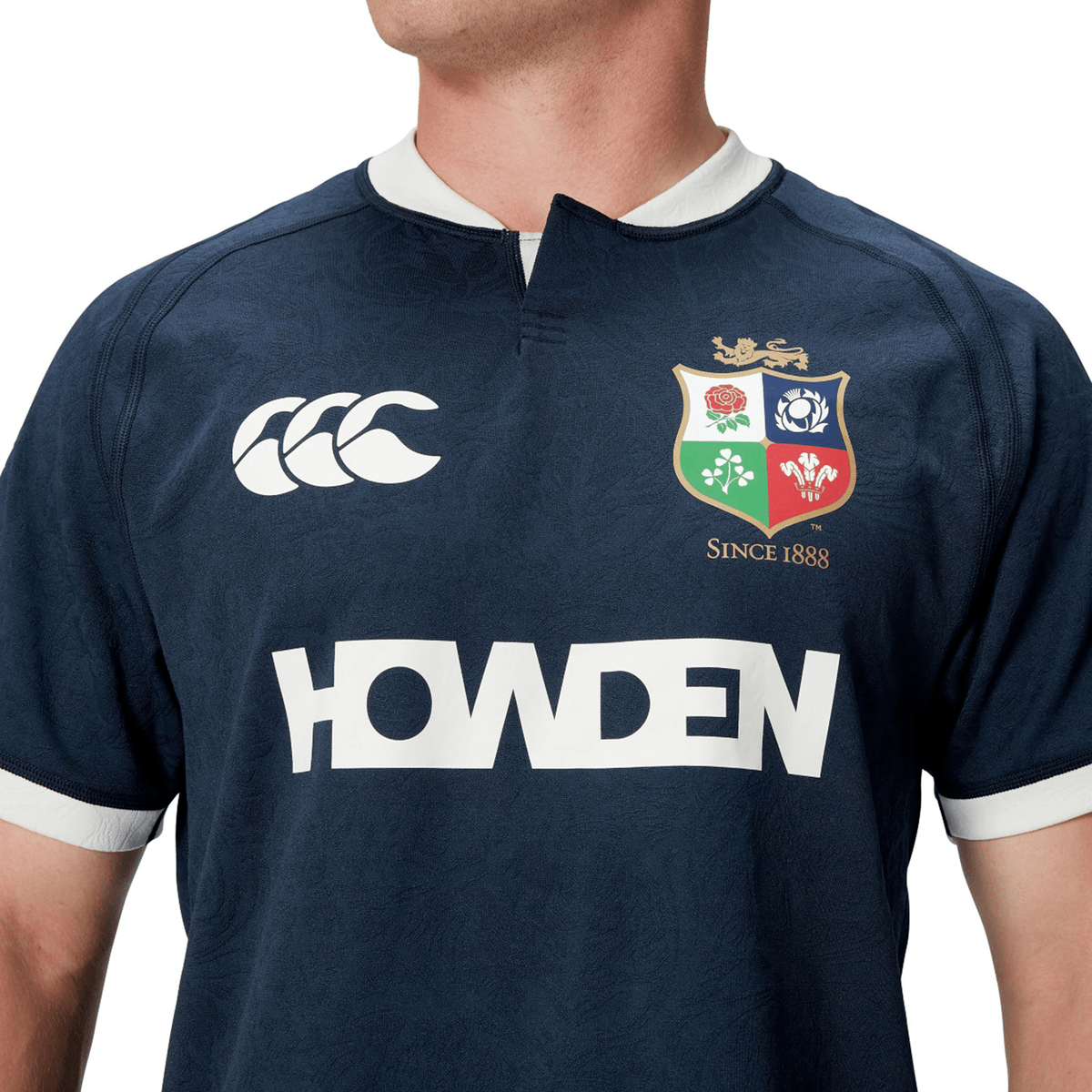 A person wearing the British &amp; Irish Lions 2025 Training Jersey by Canterbury, featuring a navy blue design with logos and the text "Howden" on the front, along with ergonomically placed panels for enhanced comfort.