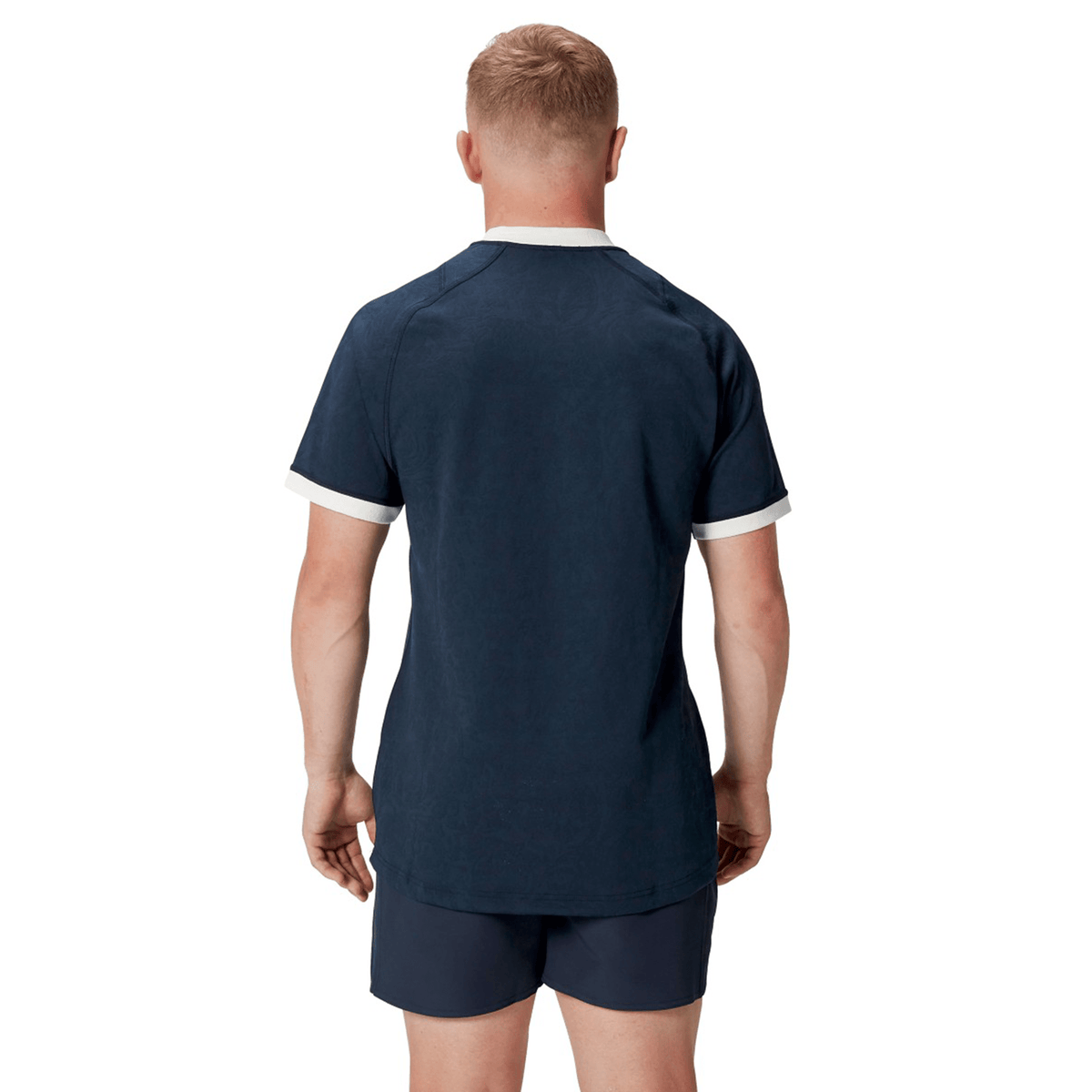A person with short blond hair is wearing the British &amp; Irish Lions 2025 Training Jersey by Canterbury, featuring ergonomically placed panels for comfort, paired with navy blue shorts, and is standing with their back to the camera.