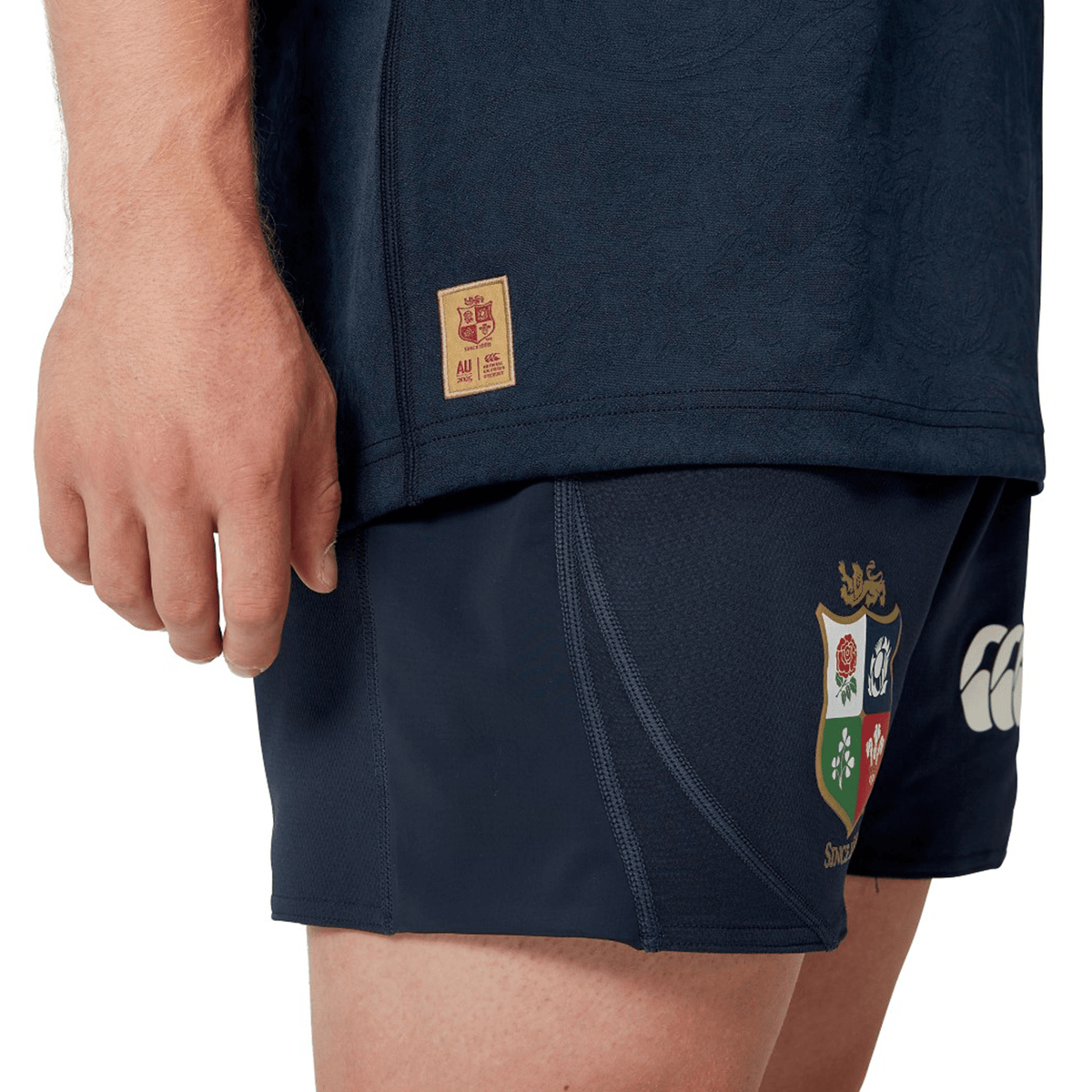 A person in a dark sports outfit is wearing the British &amp; Irish Lions 2025 Training Jersey by Canterbury, which features ergonomically placed panels, along with shorts embellished with the colorful crest and logo of the British &amp; Irish Lions.