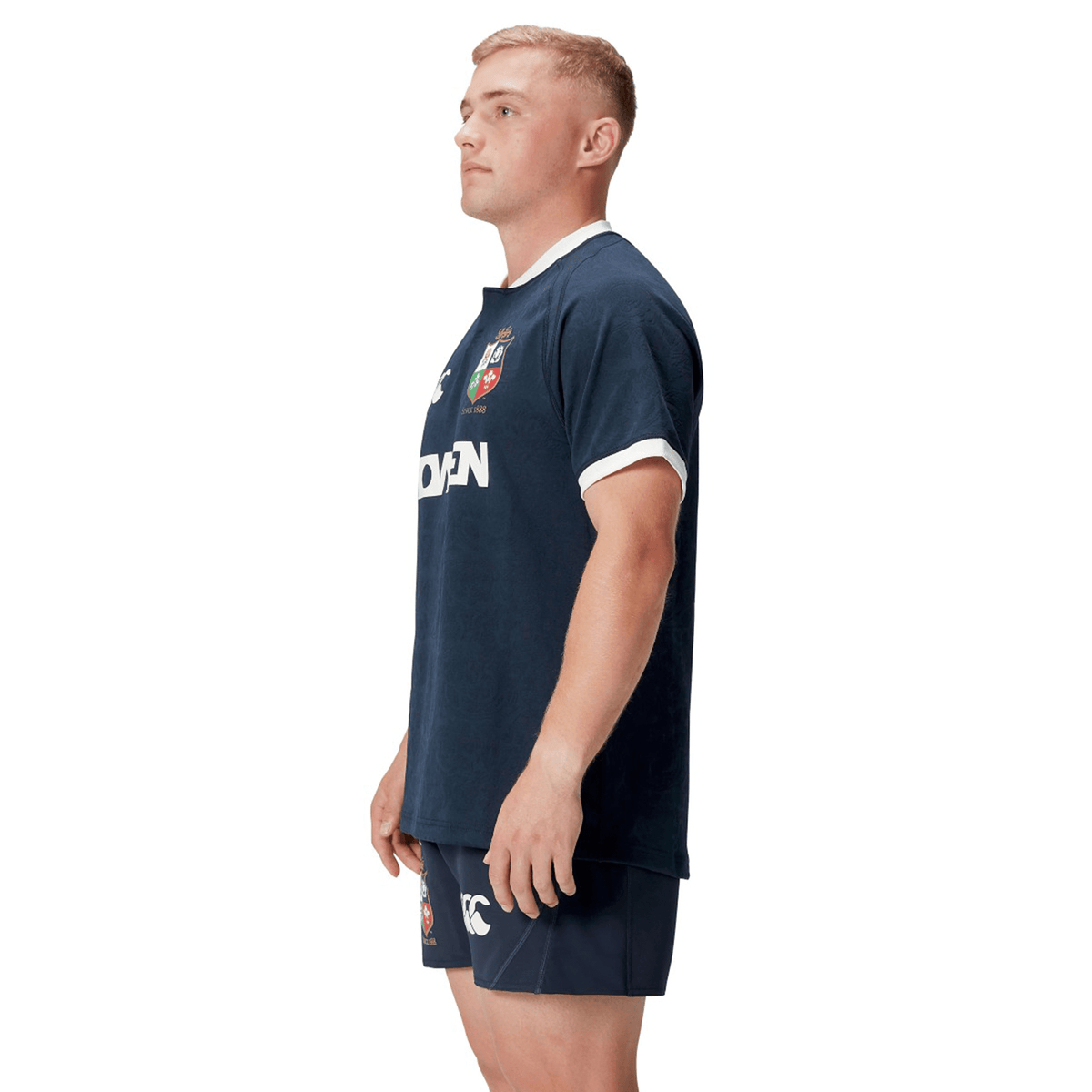 A man stands in profile wearing the British &amp; Irish Lions 2025 Training Jersey by Canterbury. This navy rugby uniform is adorned with red and white crest logos, inspired by the classic British &amp; Irish Lions design, and crafted from recycled polyester, combining style and sustainability.