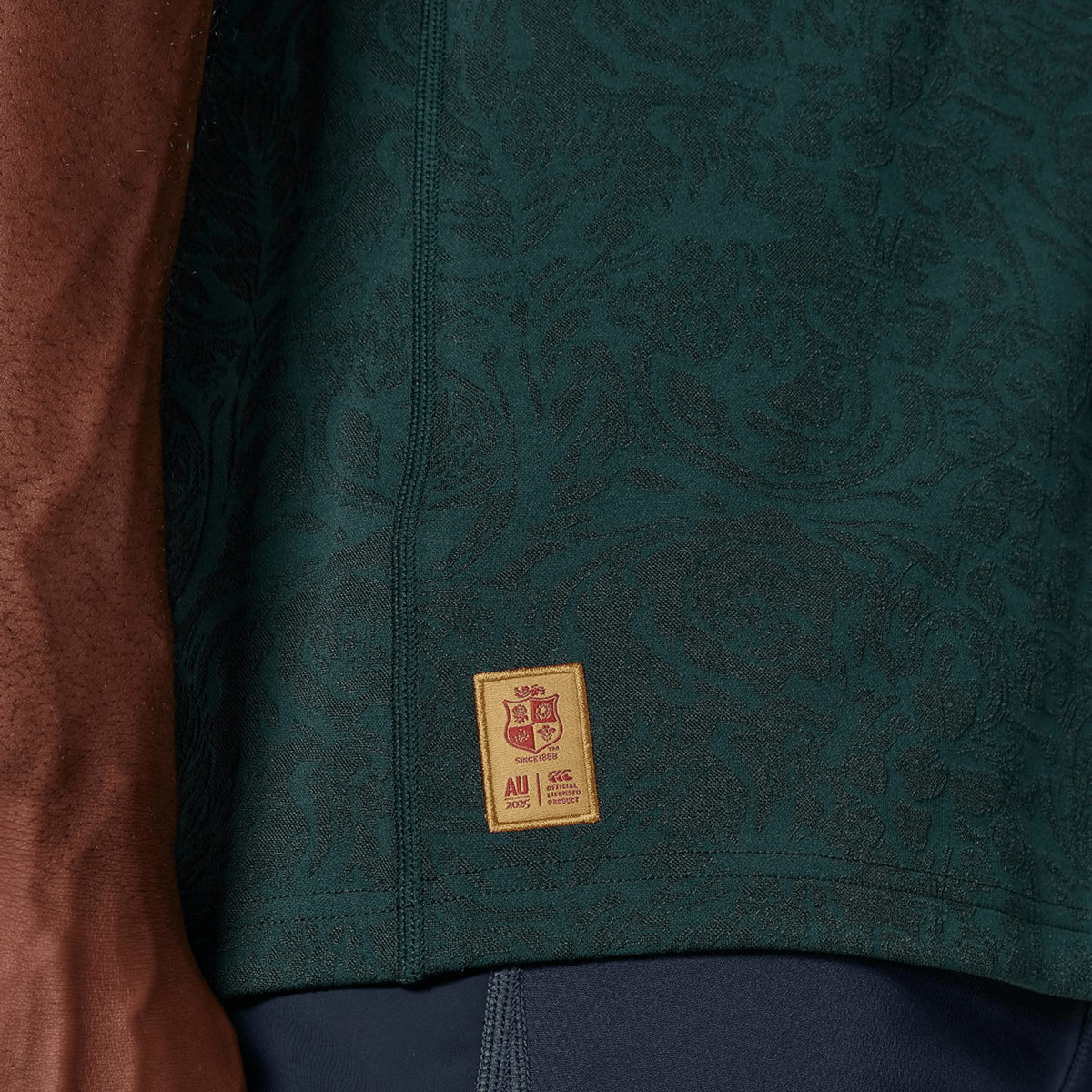 Close-up of a person wearing the British &amp; Irish Lions 2025 Training Jersey by Canterbury, showcasing a dark green pattern with a small yellow and red rectangular patch at the bottom.