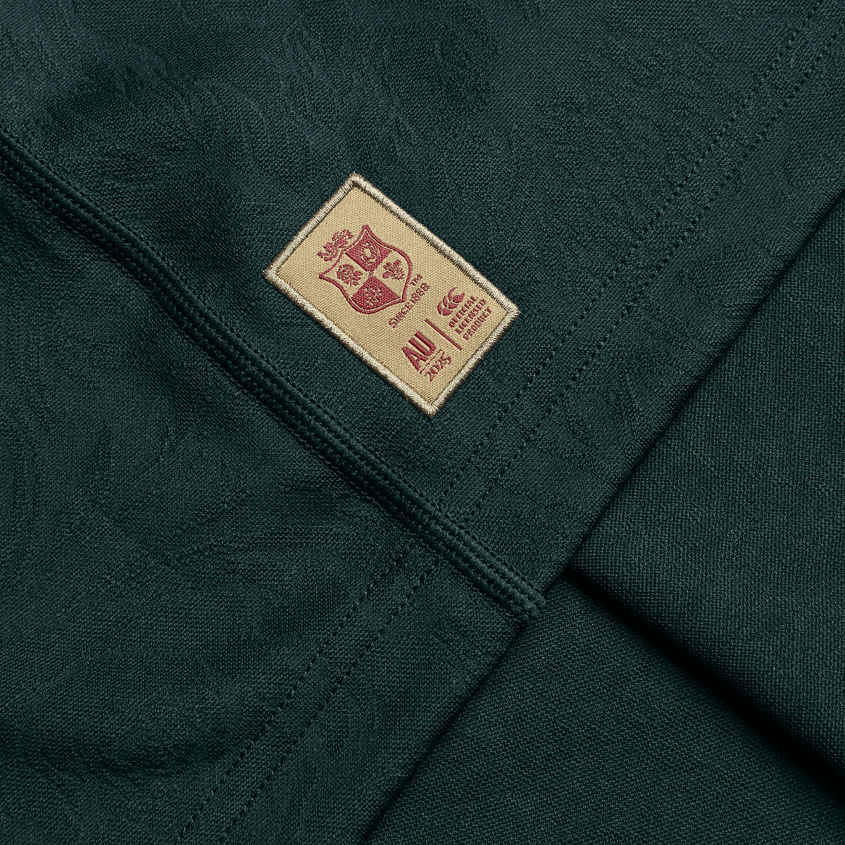 Close-up of a dark green fabric, crafted from recycled polyester, adorned with a beige patch stitched with a maroon crest and the year 2014 alongside the letters "AU," evoking the iconic style of Canterbury's British &amp; Irish Lions 2025 Training Jersey.