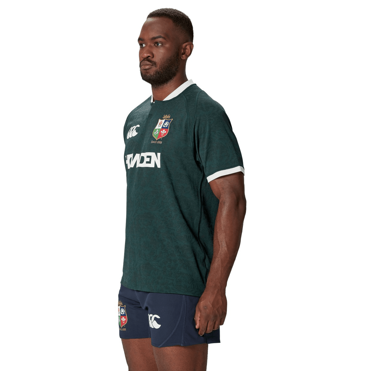 A man in a Canterbury-designed green and blue sports uniform, crafted from recycled polyester, stands facing sideways, closely resembling the British &amp; Irish Lions 2025 Training Jersey.