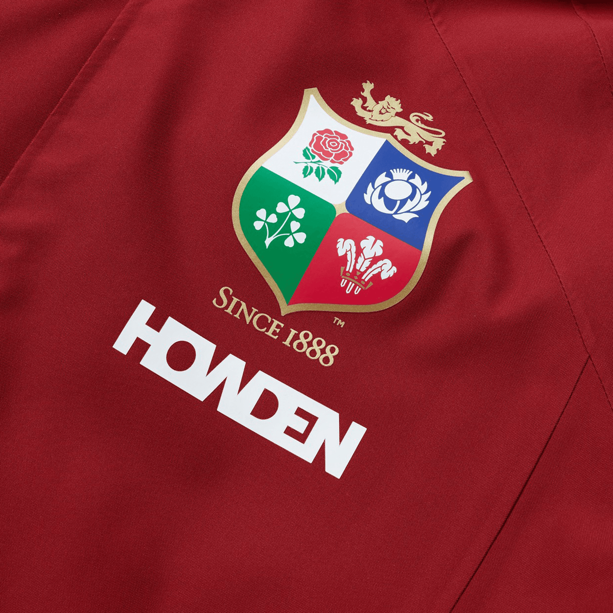 Close-up of a red polyester jacket featuring a rugby emblem with four national symbols and &quot;Since 1888&quot; above the Canterbury brand name. This waterproof jacket captures the essence of the British &amp; Irish Lions and is part of their 2025 collection by Canterbury.