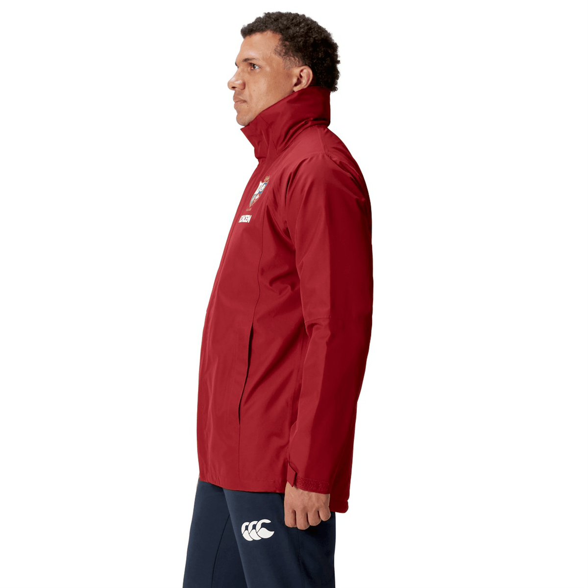 A person wearing a red British &amp; Irish Lions 2025 Waterproof Jacket by Canterbury, featuring an insulated inner layer, and dark pants stands in profile against a white background.