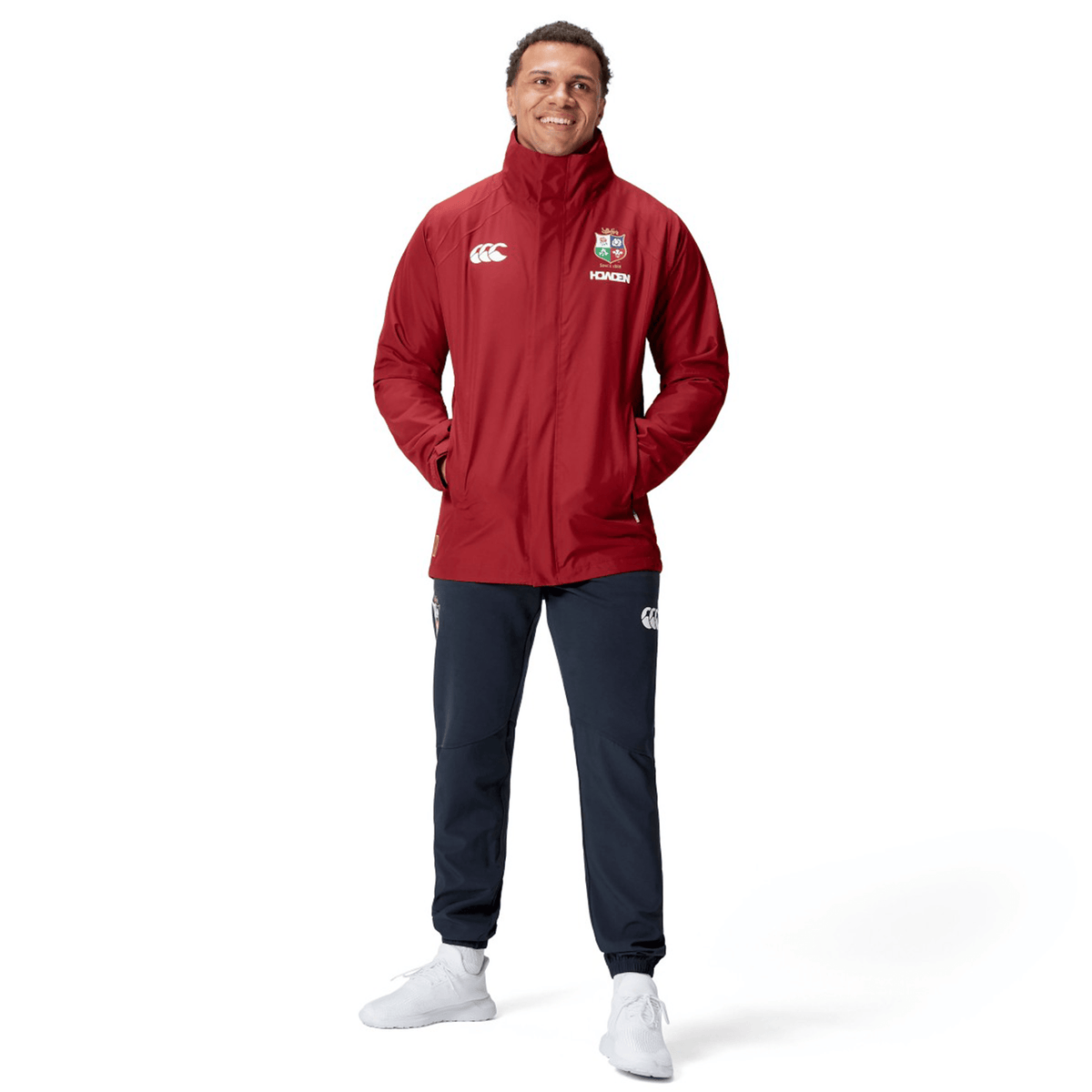 A person in a British &amp; Irish Lions 2025 Waterproof Jacket by Canterbury, featuring logos, stands smiling with hands in pockets, wearing black pants and white sneakers against a white background.