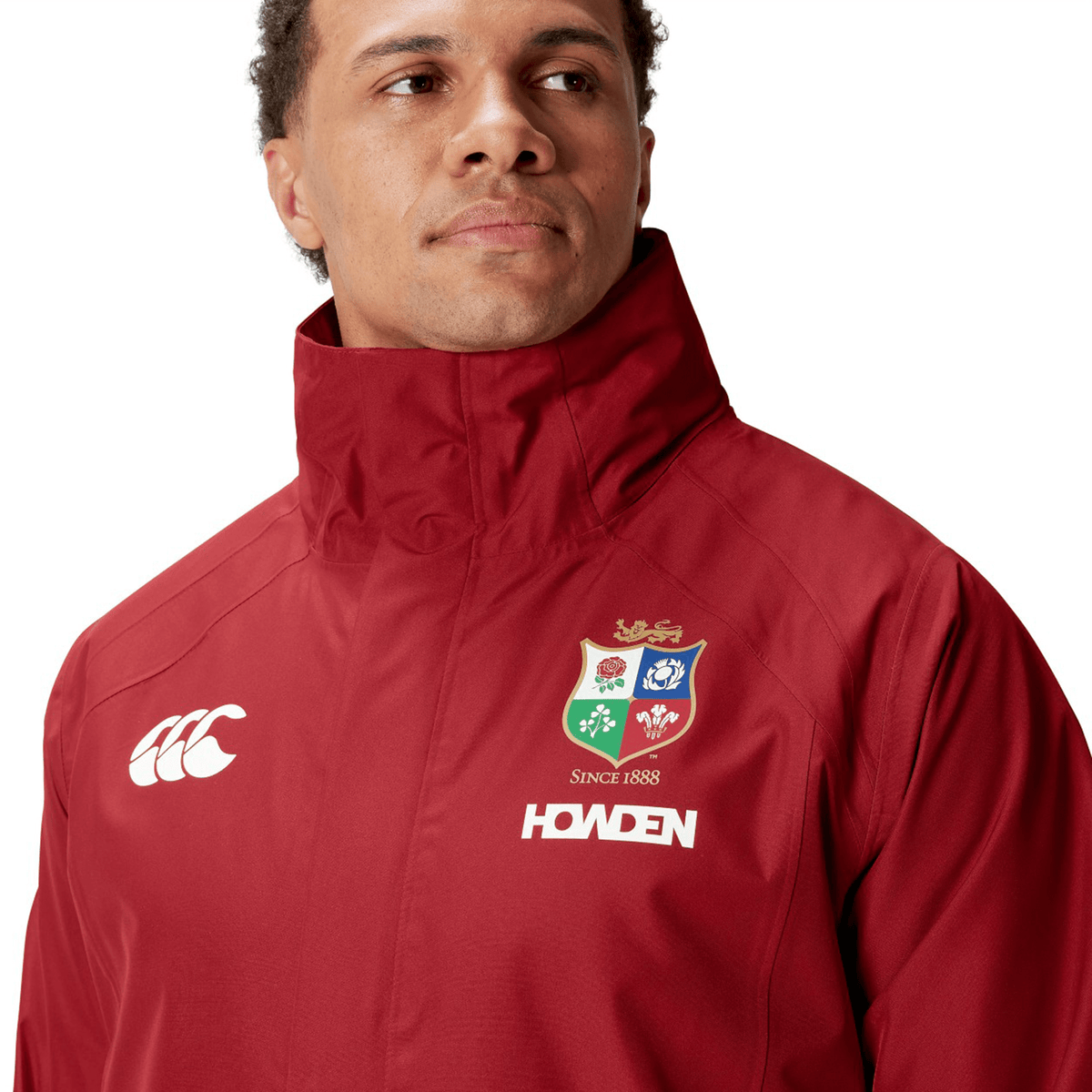 A person wearing the British &amp; Irish Lions 2025 Waterproof Jacket by Canterbury, crafted from polyester in a red hue, showcasing the lion and crest logo of the British &amp; Irish Lions with &quot;SINCE 1888&quot; text.