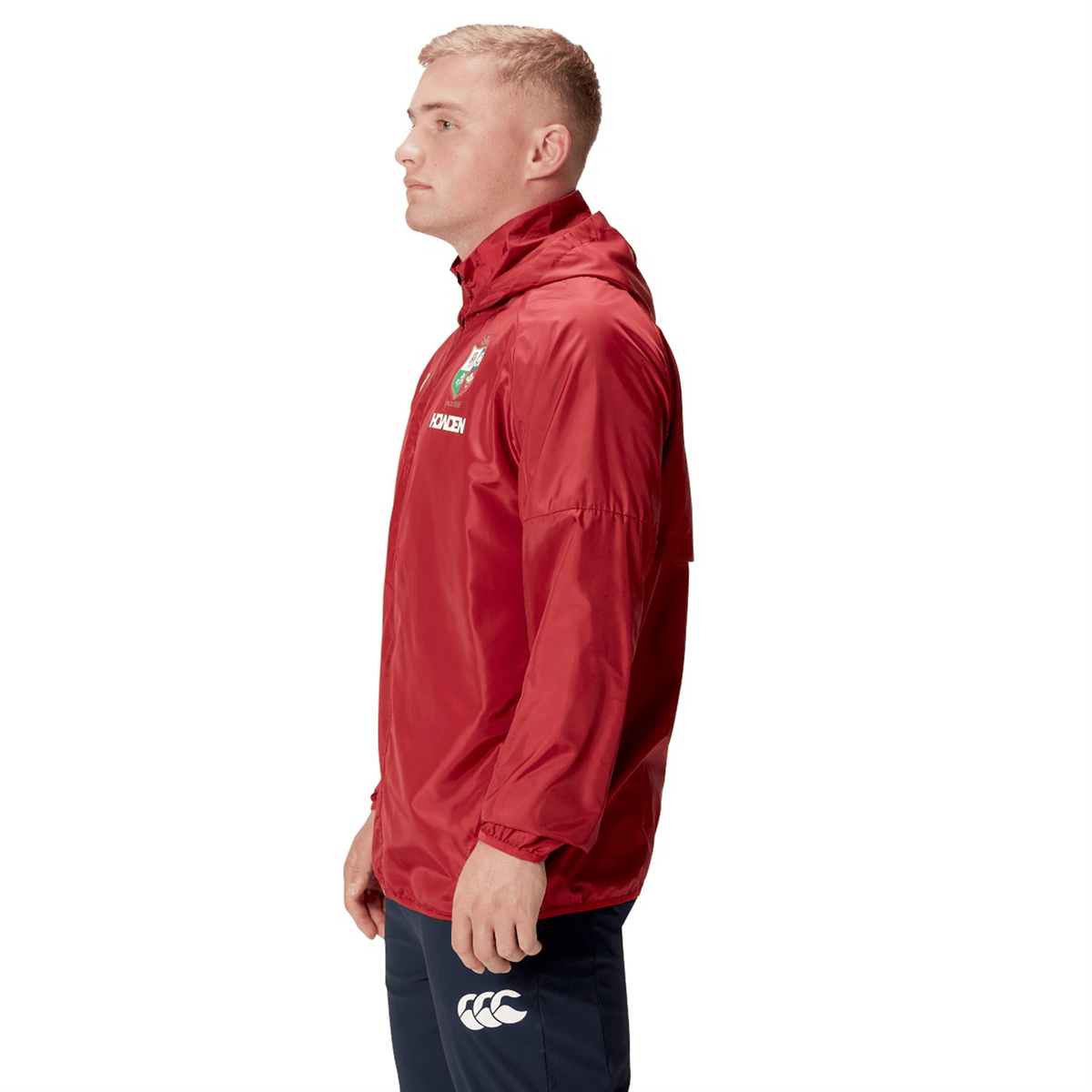 A person stands in profile, wearing the British &amp; Irish Lions 2025 Lightweight Rain Jacket by Canterbury in red with a hood, paired with navy pants featuring a logo, all set against a white background.