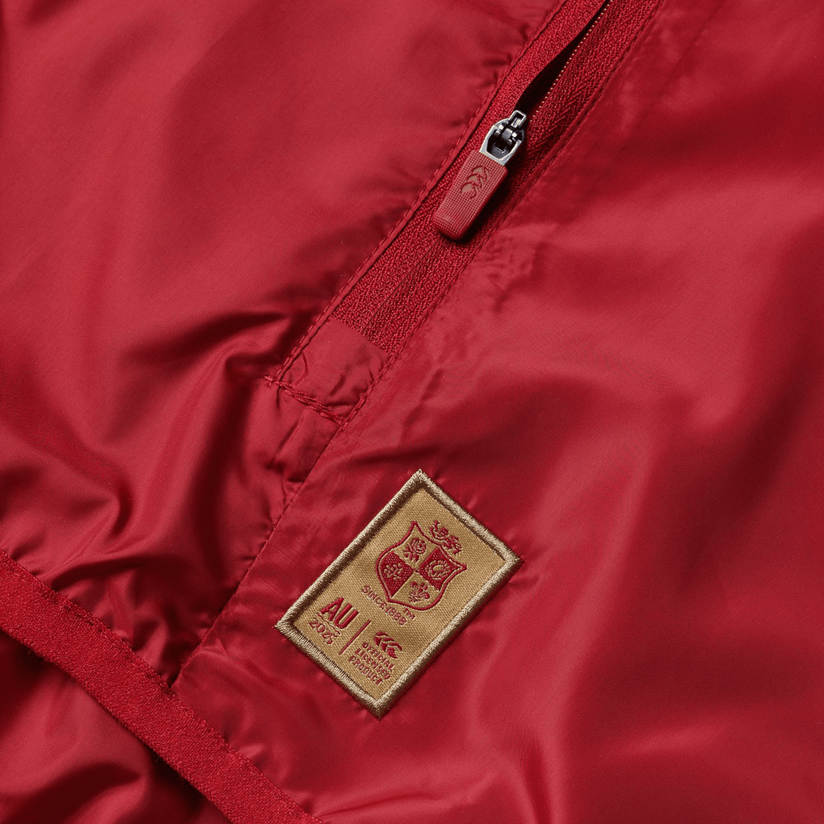 Close-up of a British &amp; Irish Lions 2025 Lightweight Rain Jacket by Canterbury, designed in red and featuring a gold patch with a coat of arms and a small zippered pocket, crafted from recycled polyester.