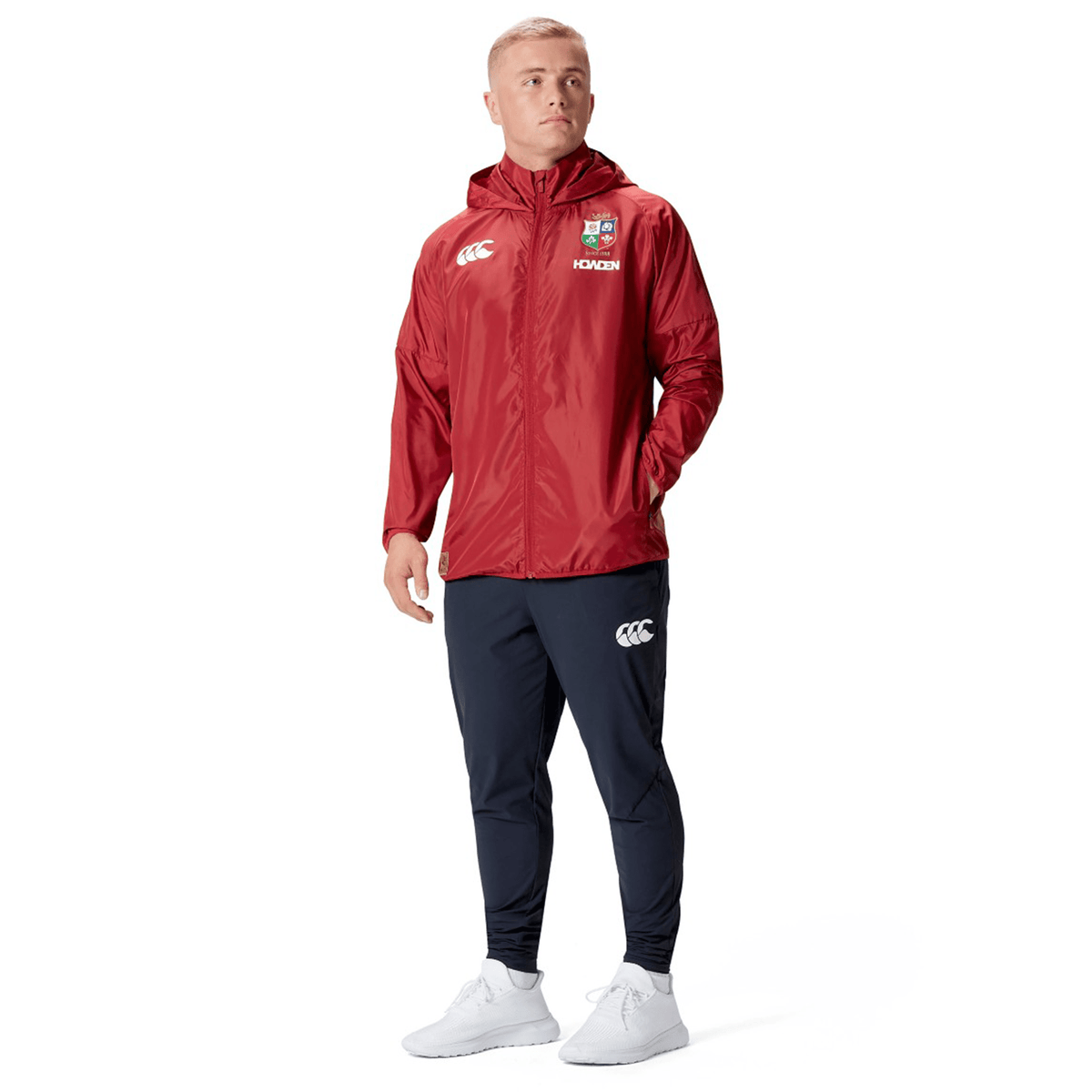 A person wearing a red British &amp; Irish Lions 2025 Lightweight Rain Jacket by Canterbury and navy blue track pants stands with hands in pockets, looking to the side.