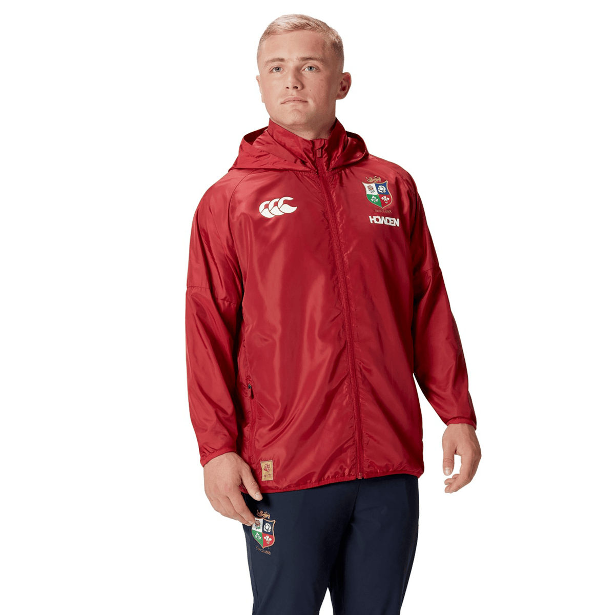 A person wearing the British &amp; Irish Lions 2025 Lightweight Rain Jacket by Canterbury, featuring logos and a red hood, stands against a white background. This stylish jacket is not only lightweight but also water-resistant, making it ideal for unpredictable weather.