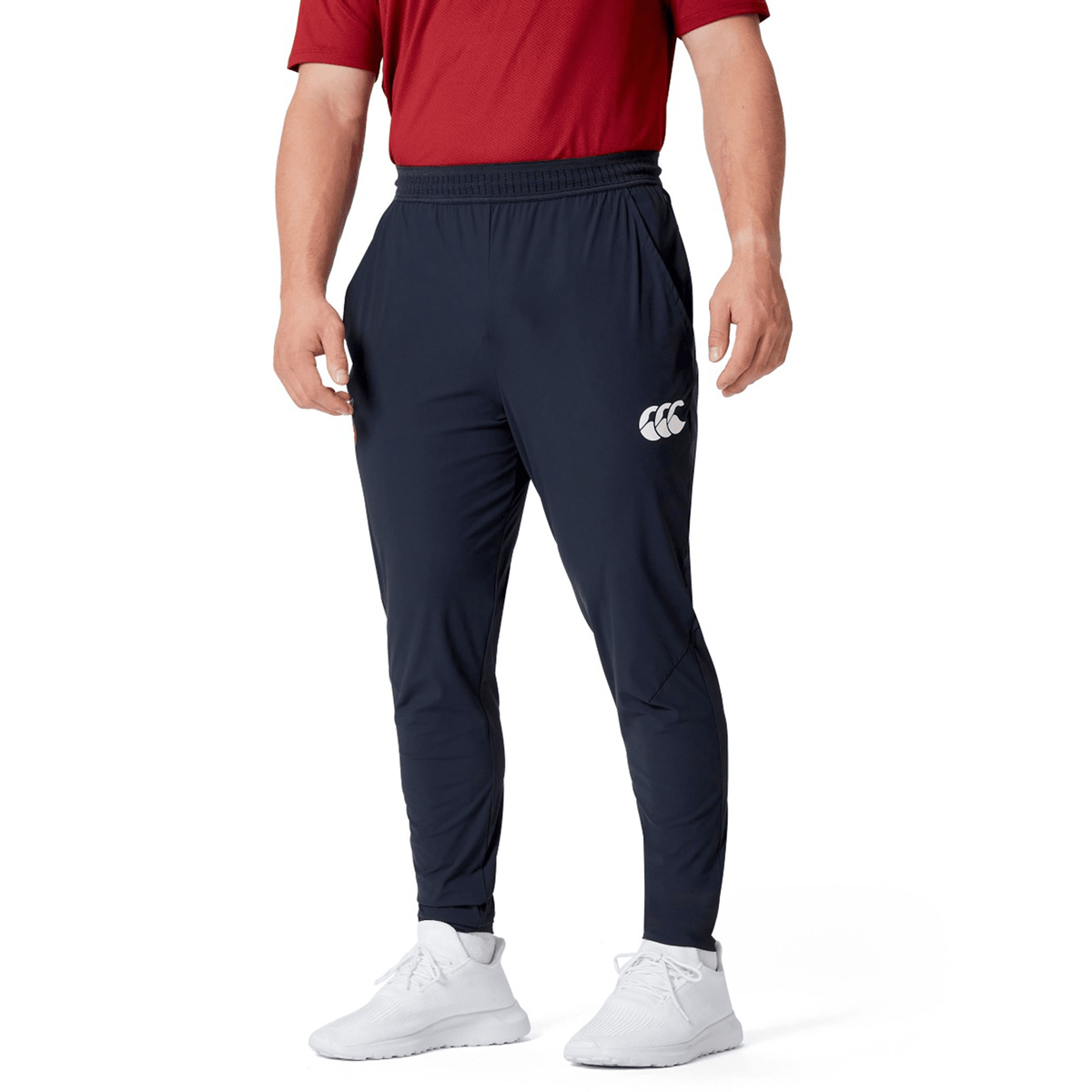 The person sports a red shirt paired with the British &amp; Irish Lions 2025 Everest Woven Pant by Canterbury, which features four-way stretch for optimum comfort. White sneakers complete the look, adding a touch of style and practicality. With hands casually tucked in pockets, they're set for quick-drying adventures.