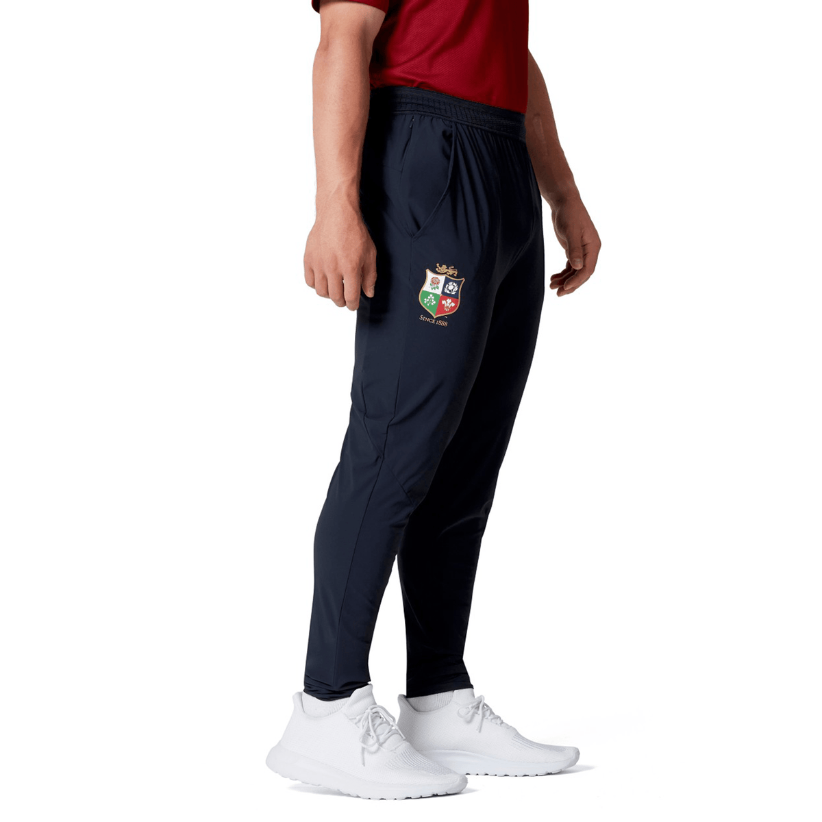 A person in a red shirt wearing the British &amp; Irish Lions 2025 Everest Woven Pant by Canterbury, featuring a crest emblem and paired with white sneakers, stands sideways. This outfit's four-way stretch fabric delivers comfort and quick-drying convenience, ideal for maintaining an active lifestyle.
