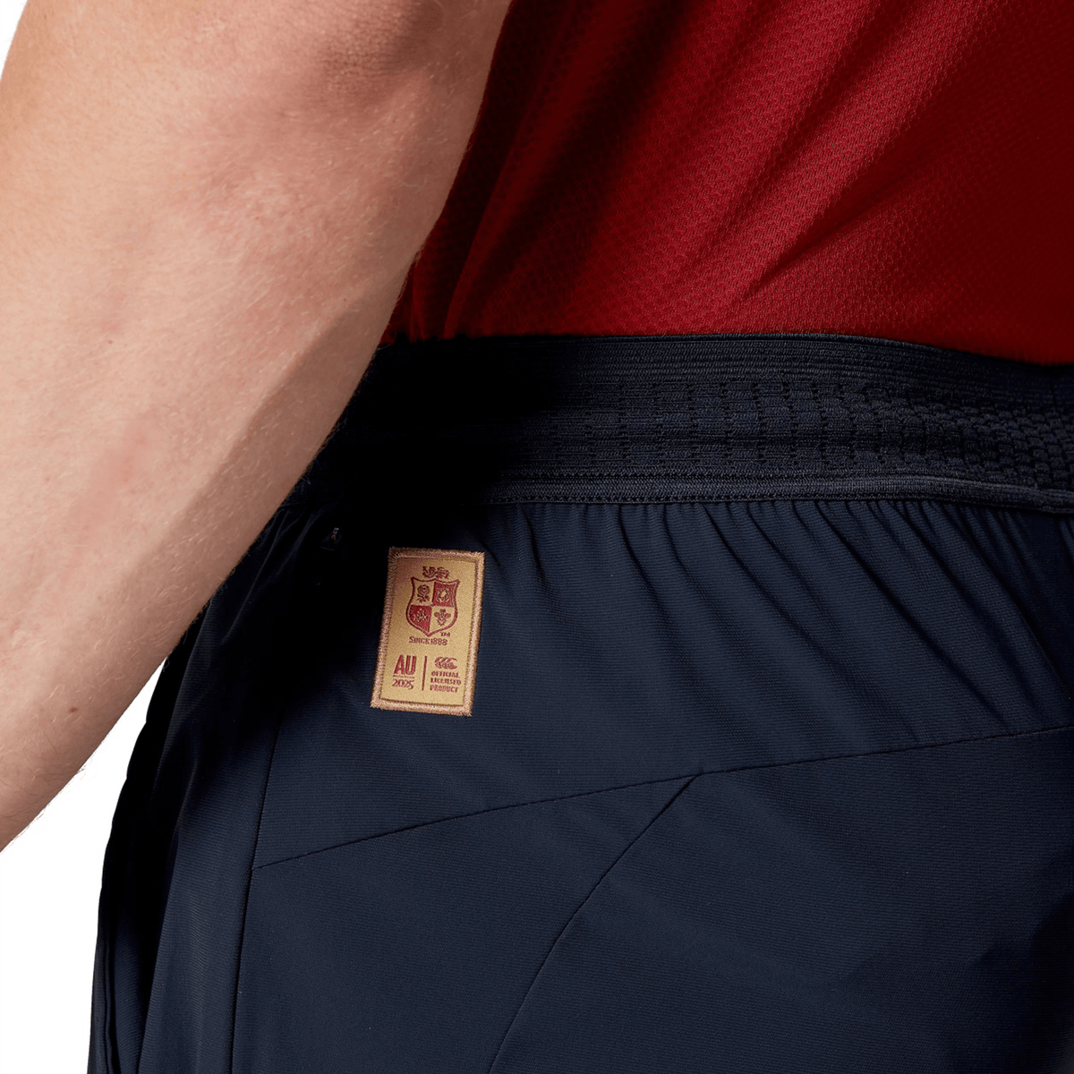 Close-up of a person donning a red shirt and the British &amp; Irish Lions 2025 Everest Woven Pant by Canterbury in navy, designed with quick-drying fabric and featuring a brown label on the waistband, perfect for channeling your inner British &amp; Irish Lions spirit.