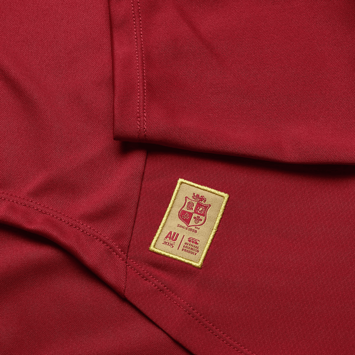 A close-up of a red sports jersey, styled like the British &amp; Irish Lions' collection from Canterbury, showcases a sewn-on gold patch adorned with a crest, "AU" text, and logos.