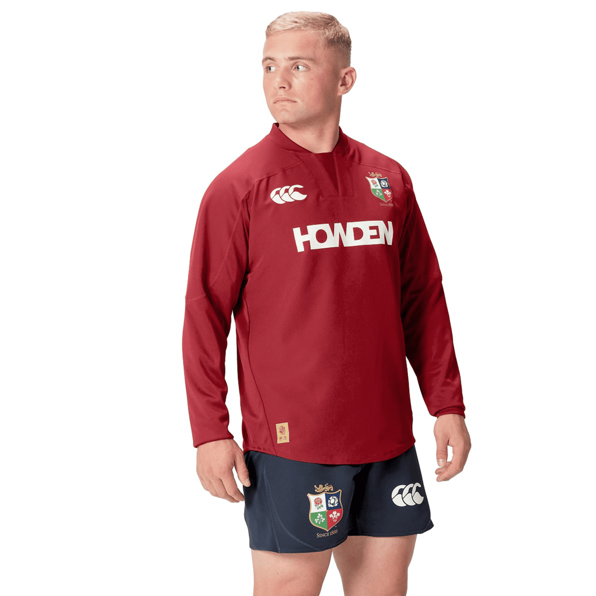 A person in a red long-sleeve British &amp; Irish Lions 25 Drill Top by Canterbury, paired with navy shorts featuring embroidered patches, stands confidently against a white background.