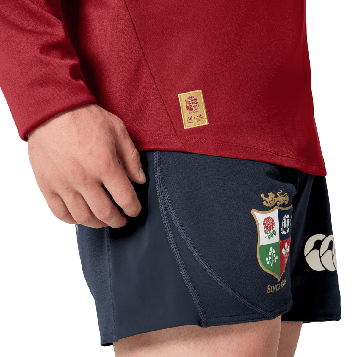A person dressed in the British &amp; Irish Lions 25 Drill Top by Canterbury, a water-resistant red long-sleeve from Canterbury, paired with dark shorts. The look is completed with a vibrant emblem adorned with floral and animal symbols, echoing the iconic style of the British &amp; Irish Lions.