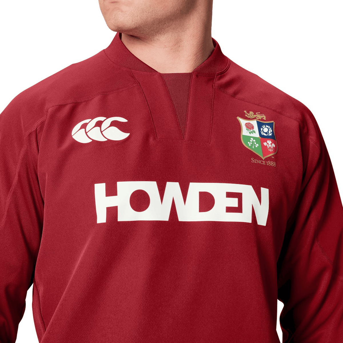 A person proudly wears the British &amp; Irish Lions 25 Drill Top by Canterbury, featuring logos like "Canterbury" and "Howden." The design is reminiscent of a durable red rugby jersey, with qualities similar to water-resistant fabric, ideal for facing the elements both on and off the field.