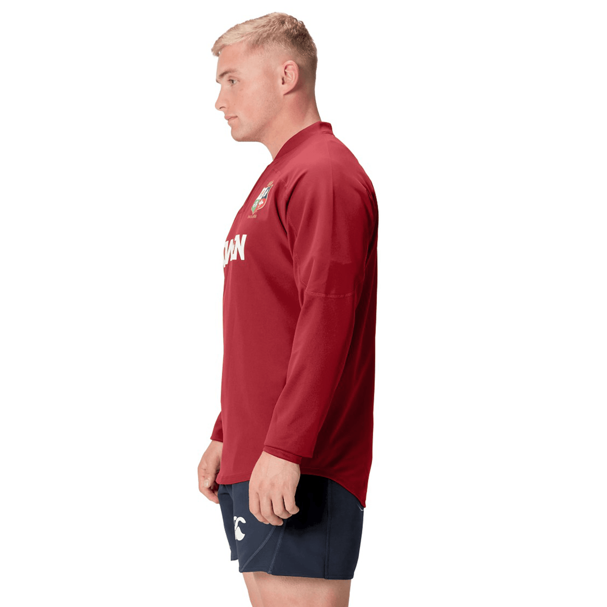A person stands in profile, wearing a long-sleeve red British &amp; Irish Lions 25 Drill Top by Canterbury and blue shorts against a white background.