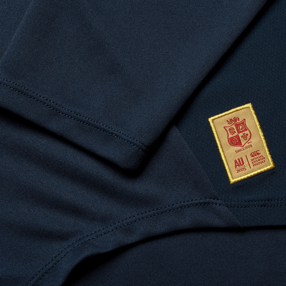 A detailed view of Canterbury's British &amp; Irish Lions 25 Drill Top showcases its water-resistant, dark blue fabric, accented by a yellow patch with red and white heraldic symbols, and text stating "AU 2025" and "CCC," evoking the classic style associated with the British &amp; Irish Lions.