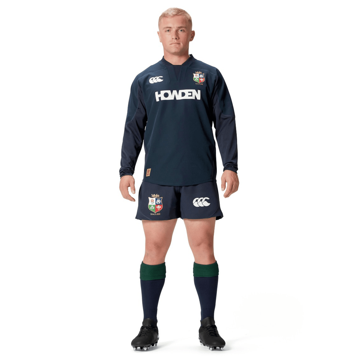 A person dressed in a navy British &amp; Irish Lions 25 Drill Top by Canterbury stands with hands by their sides, showcasing the British &amp; Irish Lions logos and black boots, all made from water-resistant fabric.