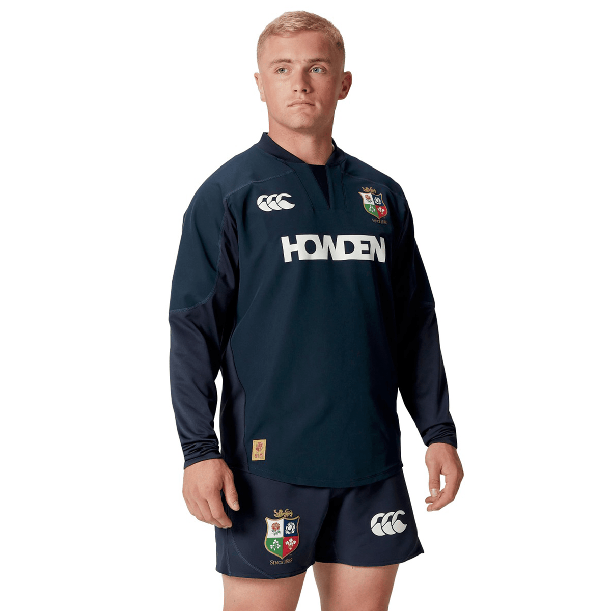 A person wearing the British &amp; Irish Lions 25 Drill Top by Canterbury, featuring sponsor logos and water-resistant fabric, stands confidently against a white background.