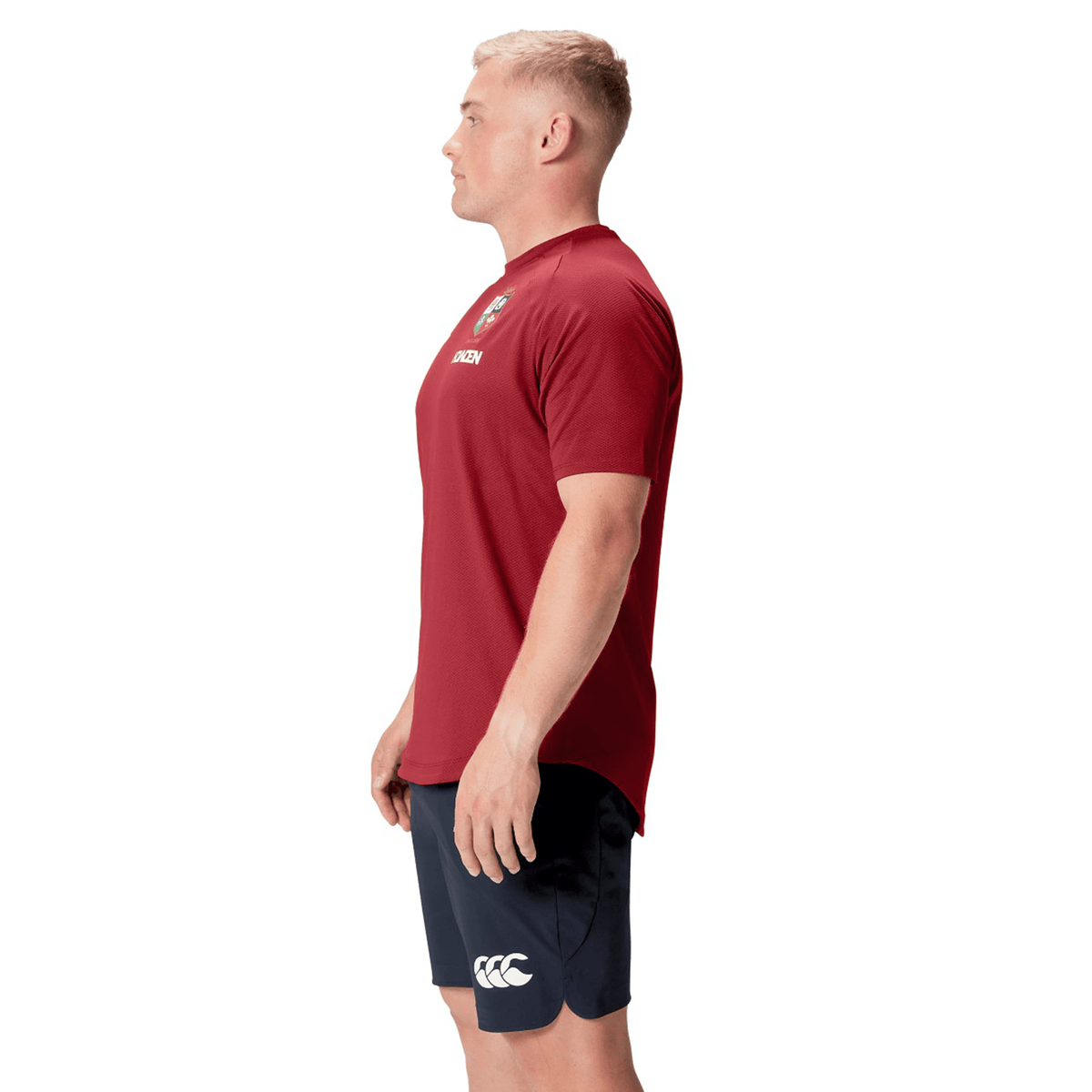 A person wearing the red British &amp; Irish Lions 25 Everest Tee by Canterbury and navy shorts stands in profile against a white background.
