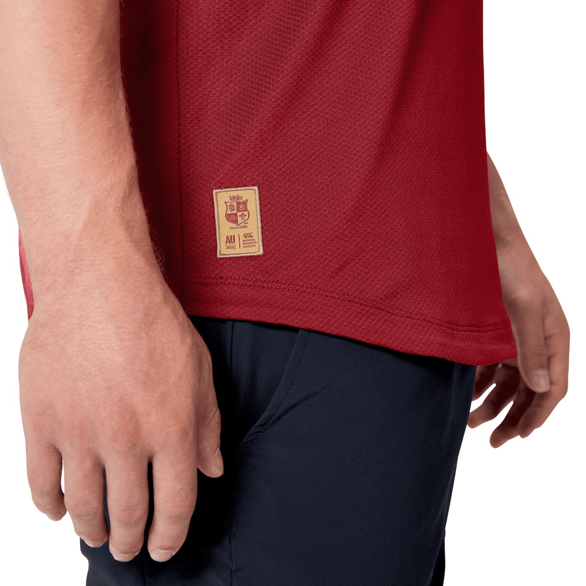 A person is wearing a vibrant red British &amp; Irish Lions 25 Everest Tee by Canterbury, complemented by black pants. This uniquely styled shirt showcases a small patch adorned with logos and text, echoing Canterbury's signature designs.