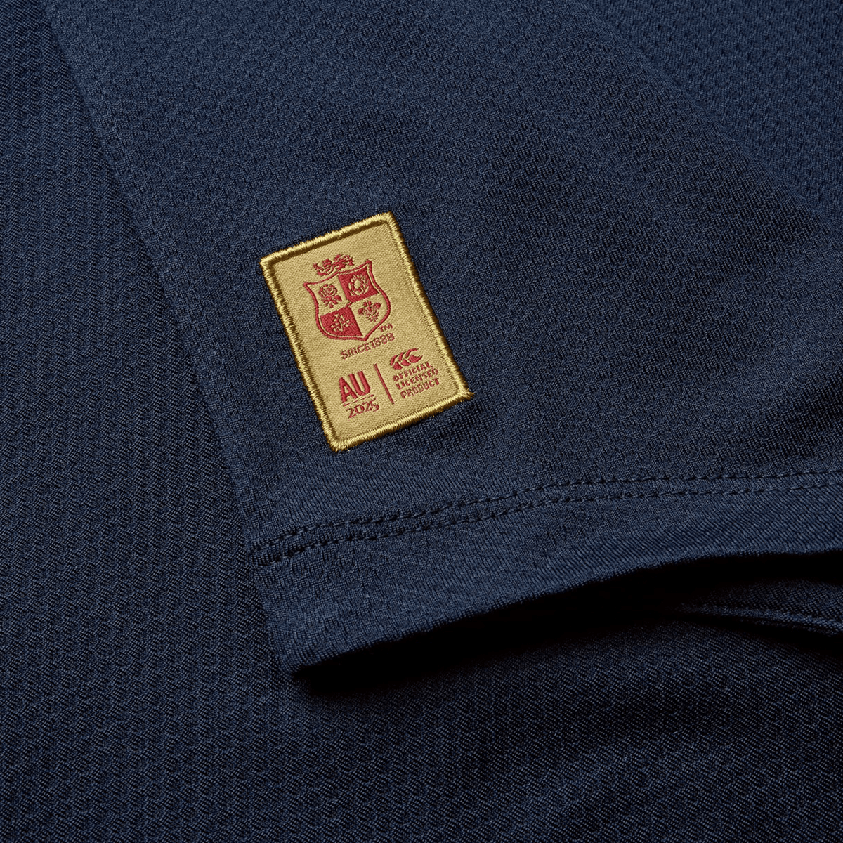 Close-up of the British &amp; Irish Lions 25 Everest Tee by Canterbury in navy, showcasing a small rectangular yellow patch with a red crest and the text "AU 2015" alongside the Canterbury logo, celebrating their British &amp; Irish Lions legacy.